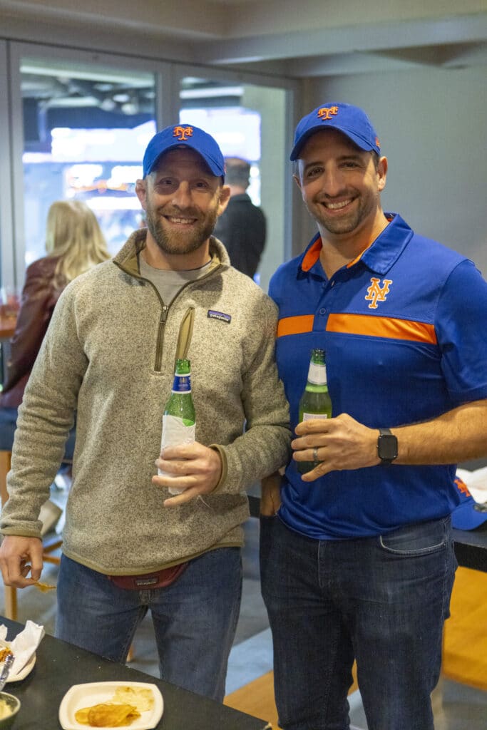 Two men posing for picture with New York Mets baseball caps on in box at game where Chopra and Nocerino threw the opening pitch