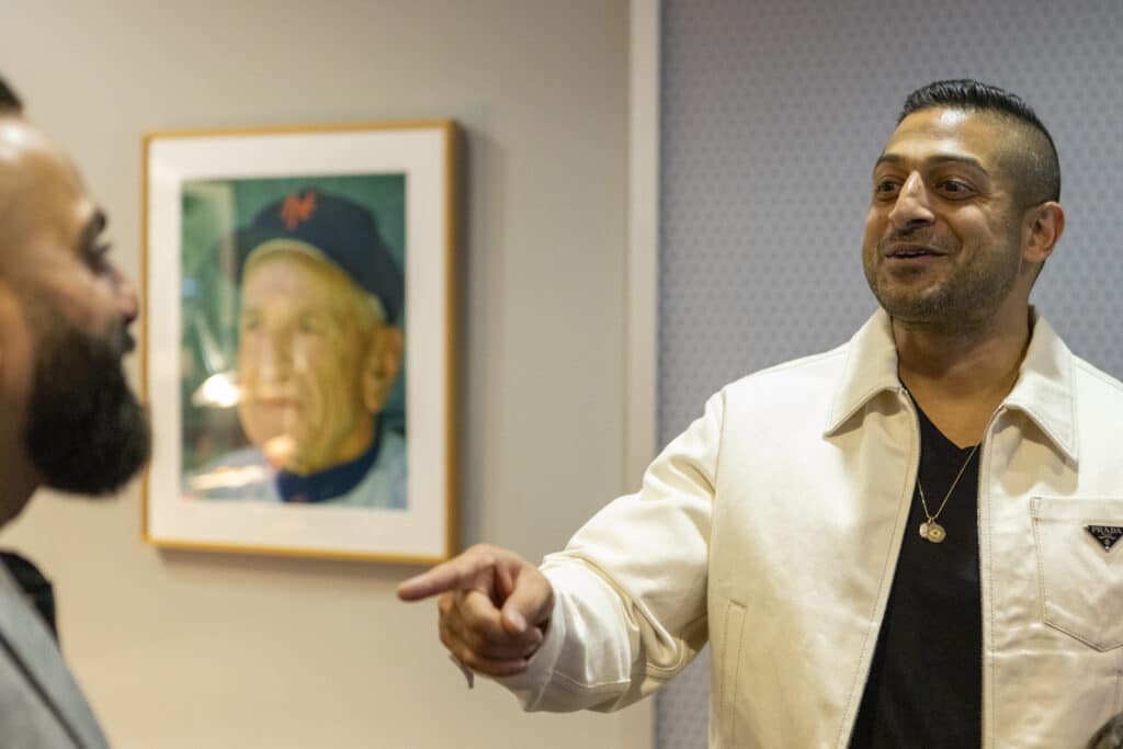 Sameer Chopra pointing and smiling at other person with New York mets portrait in background at game