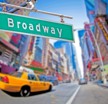 Sign of Broadway in Manhattan showcasing many colorful showsigns in background and a yellow NYC cab