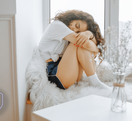 Girl in pain sitting by a window holding her knees to her chest with her eyes closed